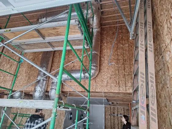 Tall insulated duct runs along OSB walls with scaffolding on a new-construction jobsite